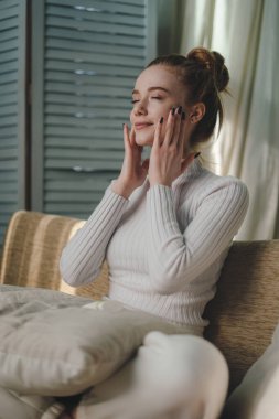 Gülümseyen kızıl saçlı kadın elleriyle yüzüne dokunuyor kusursuz cildinin tadını çıkarıyor evde güzellik ürünleriyle rutin cilt bakımı yapıyor. Evde dinleniyorum..