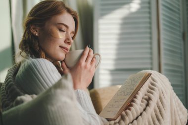 Evdeki rahat koltukta oturan, battaniyeye sarılmış, kağıt kitap okuyan, elinde fincan tutan ve sıcak kahve içen güzel sakin bir kadının portresi. Boş Zaman