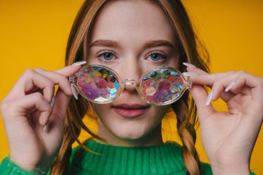 Genç, kızıl saçlı, gözlüklü bir kadının yakın plan portresi. Sarı arka planda izole edilmiş kameraya bakıyor..