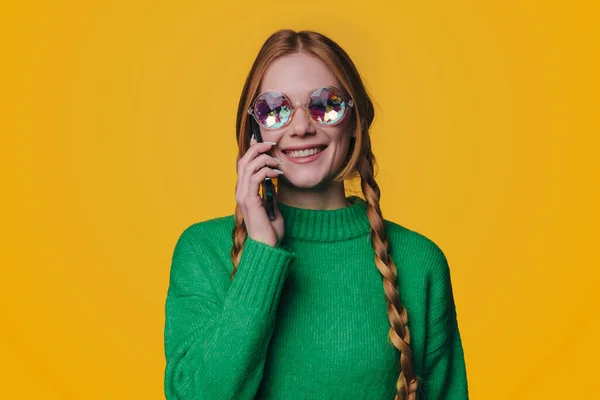 スタイリッシュな緑のセーターとサングラスを身に着けている魅力的な肯定的な笑顔若い赤髪の女性の写真黄色の背景通信上のモバイル — ストック写真