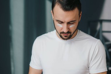 Ciddi sakallı bir adam yüzüne güzellik maskesi takıyor, banyo yaptıktan sonra sivilce tedavisi görüyor. Cilt problemi, güzellik ve hijyen kavramı.