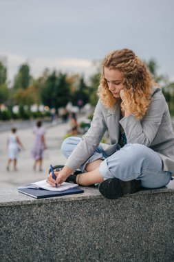 Genç ve güzel bir öğrenci parkta ders çalışıyor. Defterine yazı yazıyor. Ciddi bir şekilde konsantre olmuş, ödevlerini yapıyor. Not defterine not alıyorum.