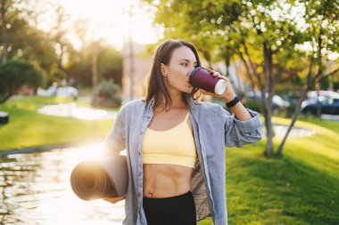 Güzel, beyaz bir kadın parkta yoga minderi tutuyor bir fincan çay ya da kahve içiyor ve başka tarafa bakıyor. Sağlıklı yaşam tarzı, sağlık. Sağlıklı yaşam tarzı, spor