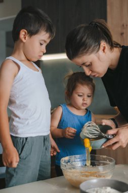 Sevimli beyaz çocuklar ve neşeli anne mutfakta birlikte yemek pişirirken kaseye yumurta koyuyorlar. Aile birlikte vakit geçirip eğleniyor..