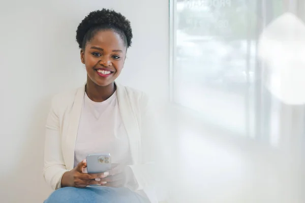 stock image Portrait of attractive young African American woman with afro hair wearing white jacket reading messages on the phone sitting in a cafe. Online communication