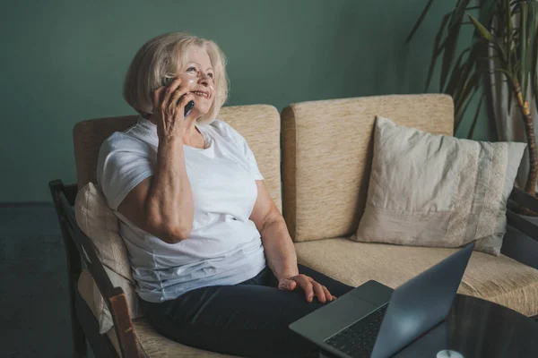 stock image Active smiling old woman talking on mobile phone while using laptop in living room at home. Woman surfing internet. Online working.