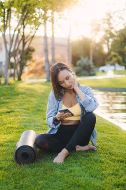 Aktif giyimli genç bir kadın çimlerin üzerinde oturuyor ve antremandan sonra cep telefonu fitness uygulaması kullanıyor. Açık havada akıllı telefon kullanan sportif bir kız. Antrenman yapıyorum..