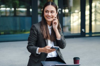 Laptopla çalışan genç ve güzel bir kadın açık havada telefon kullanıyor. Sabahları kendini iyi hissederek rahatlıyor. Kablosuz iletişim. Kadın dinleniyor. Çevrimiçi