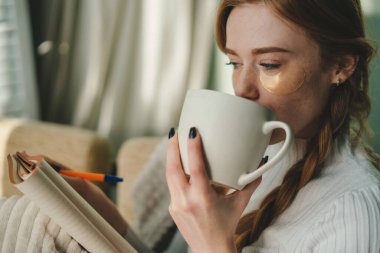 Kanepede elinde defter, bir fincan kahve olan kızıl saçlı bir kadının yumuşak fotoğrafı. Ev ofisi ya da online eğitim konsepti. Konforlu ev kavramı.