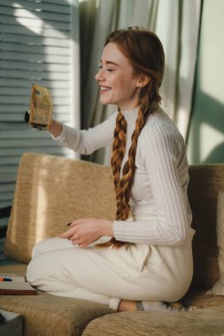 Kanepede oturan gülümseyen genç kadın not defteri ve kalem kullanıyor, not alıyor, tatil planları yapıyor, günlük tutuyor. Aile eğitimi. Yaratıcı sanat eserleri.