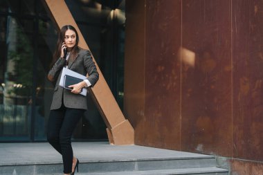 Ceketli, kendine güveni tam beyaz bir iş kadını elinde dizüstü bilgisayarla ofisin dışında müşterisiyle konuşuyor. Bina, şehir