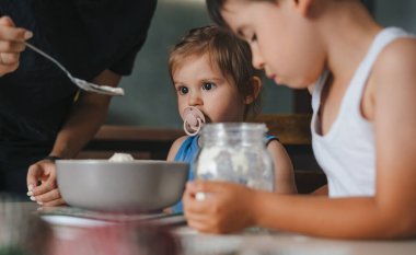 Anneler kaseye hamur malzemeleriyle un ekler, çocuklar mutfakta lezzetli hamur işleri pişirmek için annelere yardım eder..