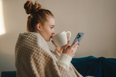 Kanepede oturup dinlenirken, sonbahar mevsiminde çay içerken akıllı telefon kullanan bir kadının yan görüntüsü. Evdeki konforlu sabah kavramı.