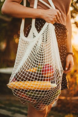 Bir kadın kol yürüyüşünün yan görüntüsü ve örülmüş sebze dolu beyaz ağ ipliği taşıyan bir alışveriş çantası. Çevre dostu, yeniden kullanılabilir alışveriş çantası. Sıfır atık