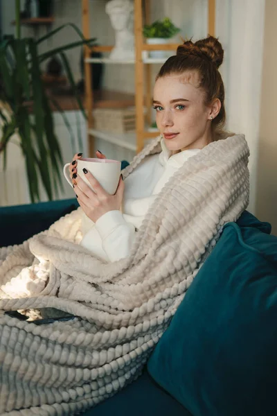 Charming positive calm peaceful woman having free time sitting on sofa with warm knitted blanket, phone and coffee. Relaxation and hygge concept. House interior