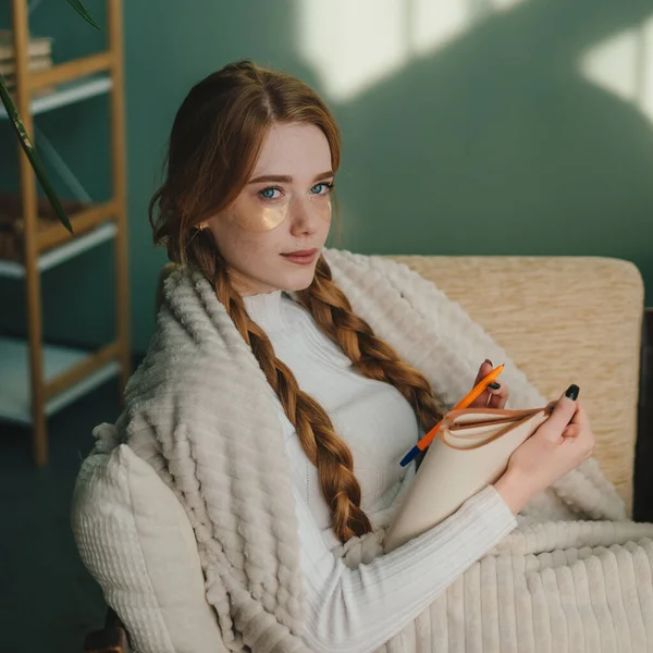 Ginger woman thinking and writing notes on a notebook, sitting on sofa covered with a blanket, looking at camera. House interior.