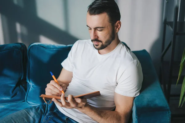 留胡子的年轻人穿着休闲装坐在客厅的沙发上 一边在笔记本上写着便条 白种人在他的日记里写日常事务 — 图库照片
