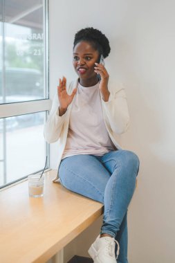 Kısa siyah kıvırcık saçlı Afro-Amerikalı bir kadın bir kafede cep telefonuyla oturuyor. Kavram: telefonu kullanmak. Cep telefonu iletişimi.