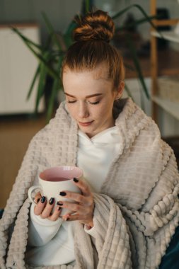 Evdeki kanepede battaniyenin altında bir fincan kahveyle oturan güzel bir kızıl kadının portresi. Tembellik konsepti. Kadın dinleniyor..