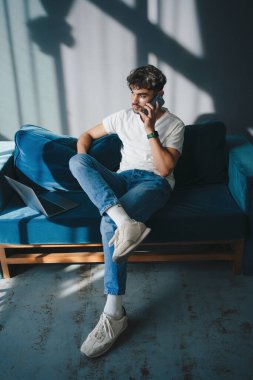 Oturma odasındaki kanepede oturan, cep telefonuyla konuşan, gündelik kıyafetler giyen çekici, sakallı bir adamın portresi. Cep telefonu iletişimi.