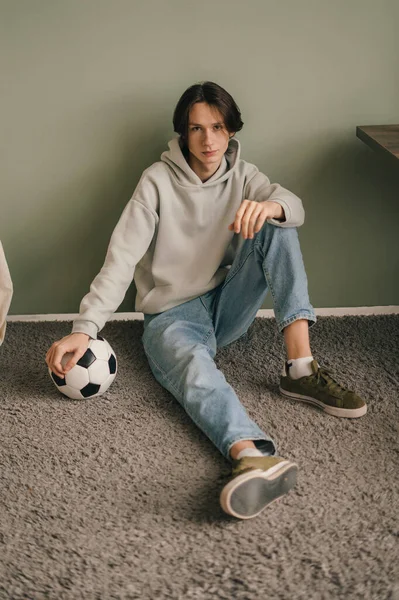 Atrakcyjny Młodzieniec Trzymający Piłkę Nożną Siedzącą Podłodze Domu Patrzącą Kamerę — Zdjęcie stockowe
