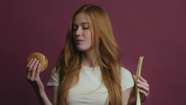 ハンバーガーを食べることを選択し 深紅の背景に隔離された野菜を選択したくない空腹の女性 健康的でないものと健康的でないもの 選択の概念 — ストック動画