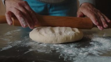 Yaşlı bir kadın elinde oklavayla un içinde hamuru uzatıyor. Fırın ve pastane kavramı.