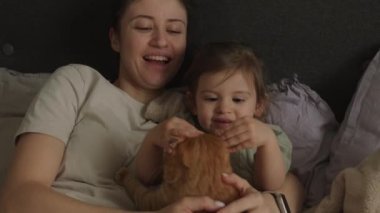 Aile grubu: Anne ve bebek yatakta uzanmış kızıl bir kediyi okşuyor. Sahibi ve evcil kedi hayvanı arasındaki ilişki. Sevimli tüylü kedi yavrusu.