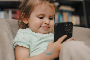 Sevimli beyaz bir kız çocuğu çizgi film izlemek için akıllı telefon kullanmaya çalışıyor, kanepede oturuyor. Bebek ve teknoloji konsepti. Mutlu bir yaşam tarzı. İnsan yaşam tarzı