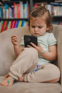 Sevimli küçük kız akıllı telefon kullanarak kanepede oturmuş oyun oynuyor. İnsanların yaşam tarzı portresi. Çocuğu oynuyorum. Eğitim interneti