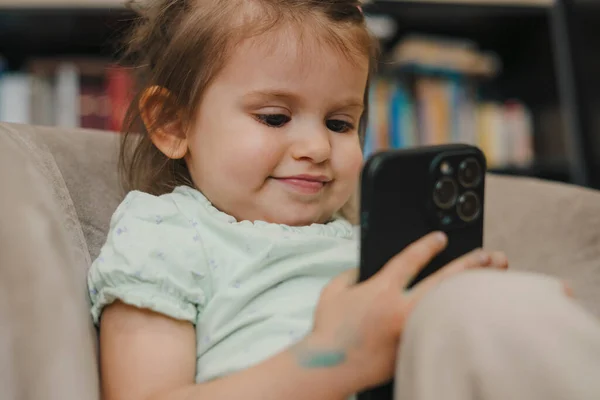 可爱的高加索小女孩坐在沙发上 试图用智能手机看卡通片 婴儿和技术概念 快乐的生活人的生活方式 — 图库照片