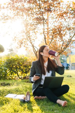 Çıplak ayaklı bir iş kadınının elinde dizüstü bilgisayarla şehir çayırında oturmuş işten sonra kahve içerken geniş açılı görüntüsü. Başarılı kadın girişimci.