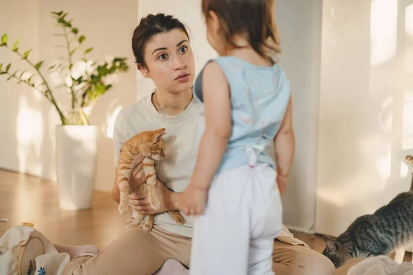 Mother and daughter playing on the floor in living room, holding pet cat. Parent and children play together. Happy young family with kid at home.