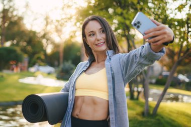 Spor kıyafetleri giyen yetişkin bir kadın spor yaptıktan sonra parkta selfie çekiyor, gülümsüyor ve spor paspası tutuyor. Sağlıklı yaşam tarzı kavramı. Eğitim