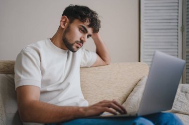 Kanepede dizüstü bilgisayarla oturan neşeli genç adam bilgisayar ekranına bakıyor. Yakışıklı genç adam sosyal medyada video izlemekten hoşlanıyor