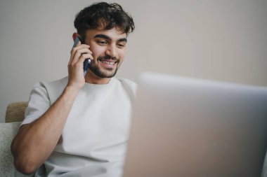 Beyaz bir adam telefon görüşmesi yapıyor, dizüstü bilgisayarla yazı yazıyor, oturma odasındaki kanepede oturuyor. Telekomünikasyon ve telgrafçılık. İletişim Kavramı.