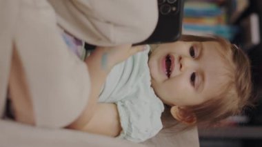 Evdeki kanepede oturan ve cep telefonuyla oynayan sevimli küçük bir kız bebek. İnternet iletişimi. Eğitim, internet teknolojisi. Çevrimiçi