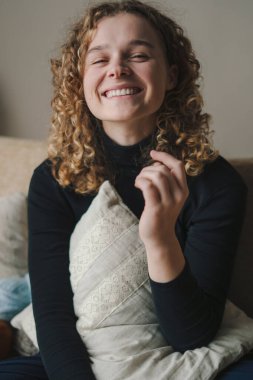 Kanepede oturan kıvırcık saçlı kadın kameraya bakıyor ve uzun dişli, sarılmış yastıkla gülümsüyor, mutlu tatiller. İnsan yaşam tarzı