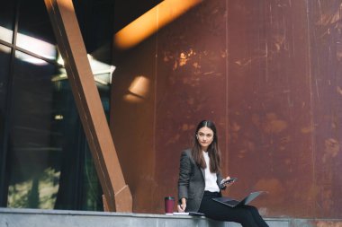 Ceketli genç ciddi girişimci akıllı telefon kullanıyor, müşterilerden gelen mesajları okuyor, çevrimiçi çalışırken dizüstü bilgisayar kullanıyor, dışarıda merdivenlerde oturuyor.