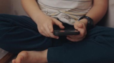 Evdeki rahat koltukta oturan çocuk elinde telefonla oyun oynuyor. Kafkasyalı sağlıklı anaokulu öğrencisi evde. Çocukluk, kumar. Çocuğu oynuyorum. Çevrimiçi iletişim
