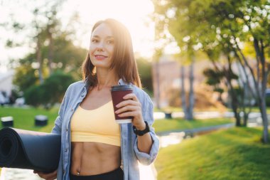 Bahçede fincanı tutan, sıcak çay içip gülümseyen, halka açık parkta dinlenen yoga minderi taşıyan çekici beyaz bir kadın. Antrenman egzersizi. İnsan vücudu