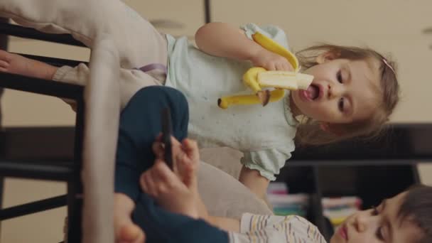 Adorável Menina Comendo Banana Olhando Para Seus Irmãos Telefone Jogar — Vídeo de Stock