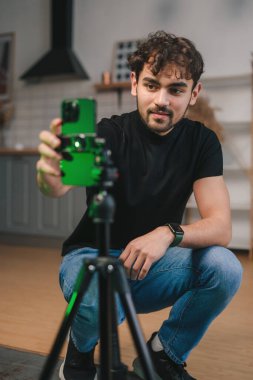 Mutlu genç adam evde video kaydediyor, sosyal ağları için içerik yaratıyor, oturma odasında tripodun üzerinde akıllı telefon kullanıyor. Ev hayatı işte. Çevrimiçi