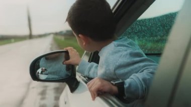 Küçük beyaz bir oğlan ve kız arabanın camından dışarı bakıyor. İnsanlar yazın tadını çıkarıyor.