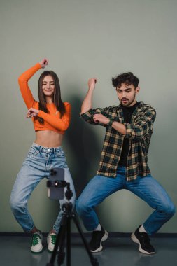 Kareli gömlekli ve kot pantolonlu, kollarını kaldırıp kameraya bakan genç bir kadın ve erkek.