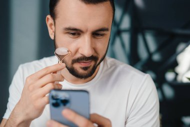 Kafede otururken, beyaz gömlekli, cep telefonunu karıştıran, konsantre sakallı bir erkek.