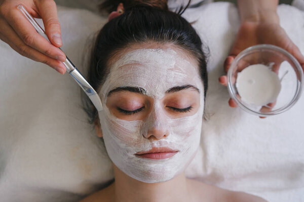 Top view of crop anonymous female applying facial mask on face with brush while sitting in light room