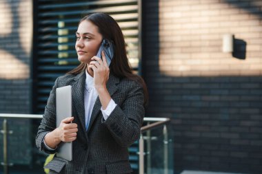 Resmi görevdeki genç iş kadını modern ofiste cep telefonuyla konuşurken gülümseyip kameraya bakıyor.