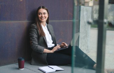 Gündelik giysiler içinde, masada dizüstü bilgisayar ve defterle otururken gülümseyen ve kameraya bakan pozitif genç bir bayan serbest yazar.