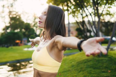 Spor kıyafetleri içinde genç bir bayanın kollarını açması ve bulanık park arka planında dikilip bakışlarını kaçırması.